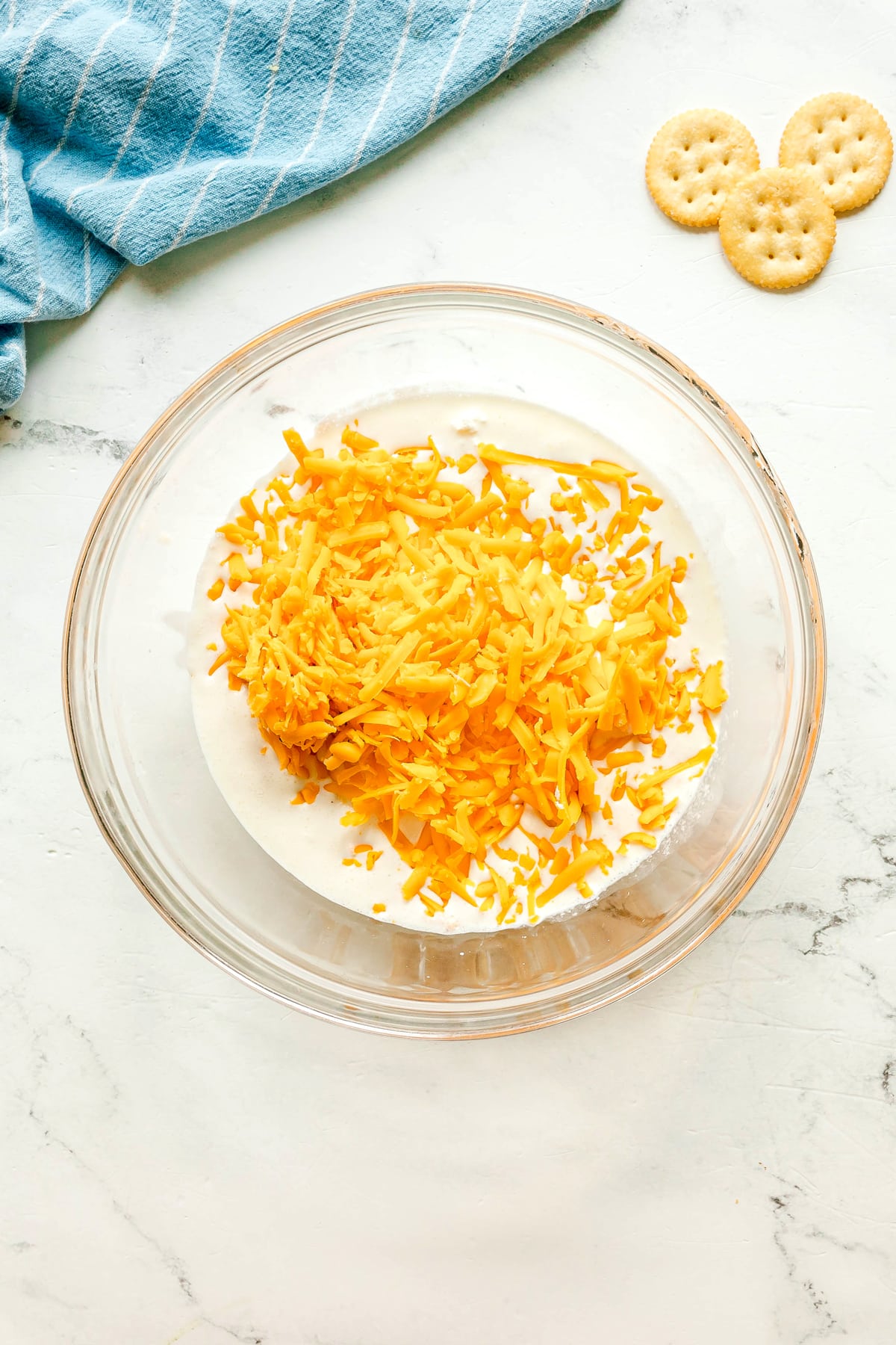 Next step in preparing Ritz Cracker Casserole is to mix cream chesse, heavy cream, chicken stock, Dijon mustard, onion powder, black pepper, and cheddar until smooth, round crackers and a blue striped towel can also be seen.