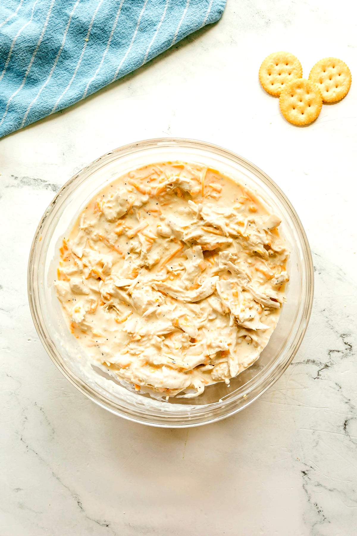 Next step in preparing Ritz Cracker Casserole is to add shredded chicken on the mixture. Three round crackers and a blue striped cloth nearby.