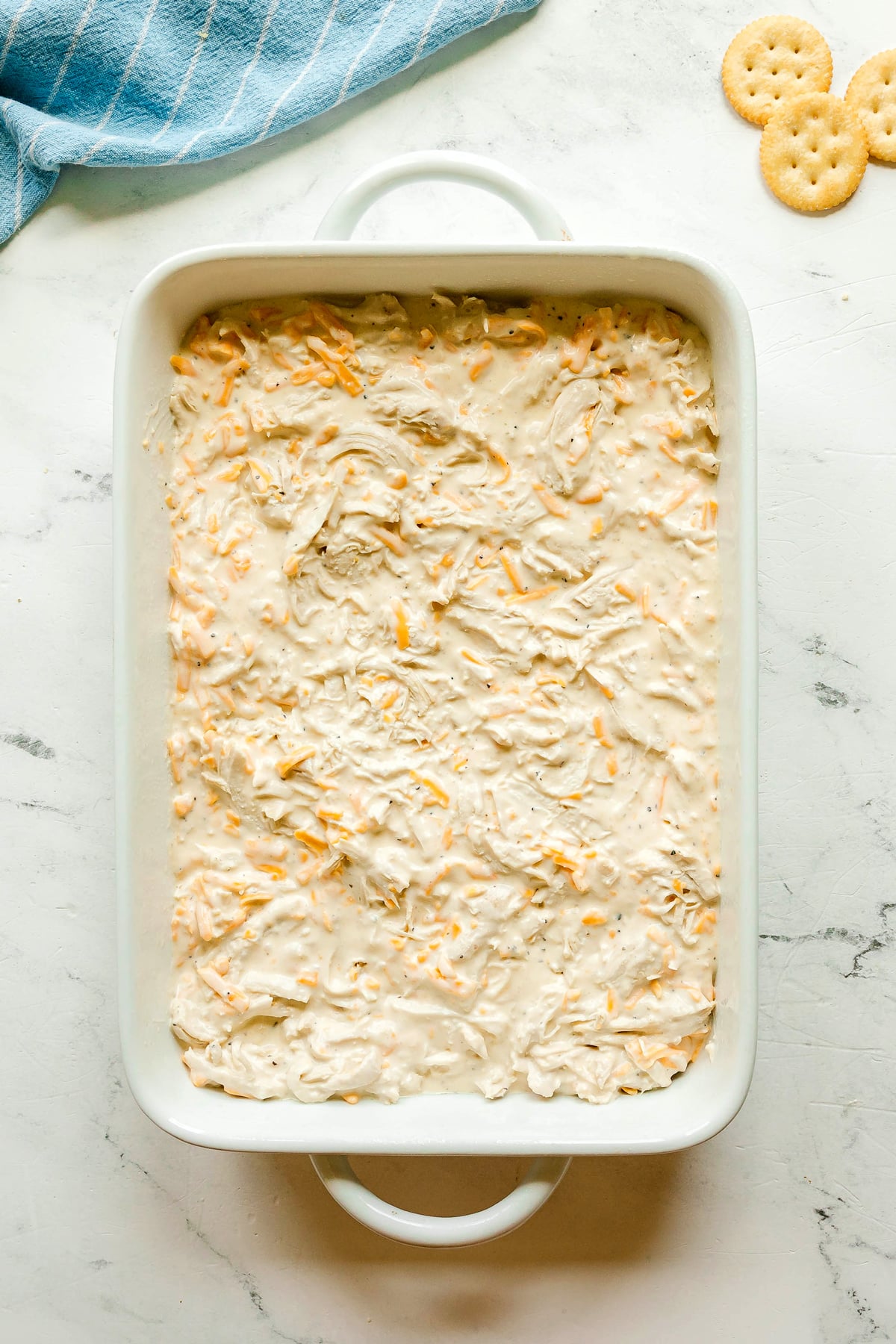 Rectangular white baking dish filled with a creamy, shredded chicken and cheese mixture of Ritz Cracker Casserole, on a marble surface; crackers and a blue towel nearby.
