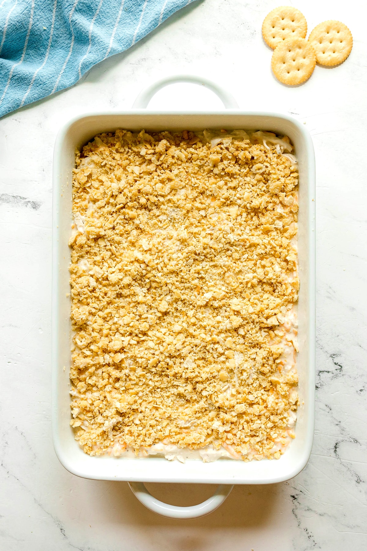 A white baking dish filled with Ritz Cracker Casserole topped with crushed crackers, placed on a white surface next to a blue striped towel and three round crackers.