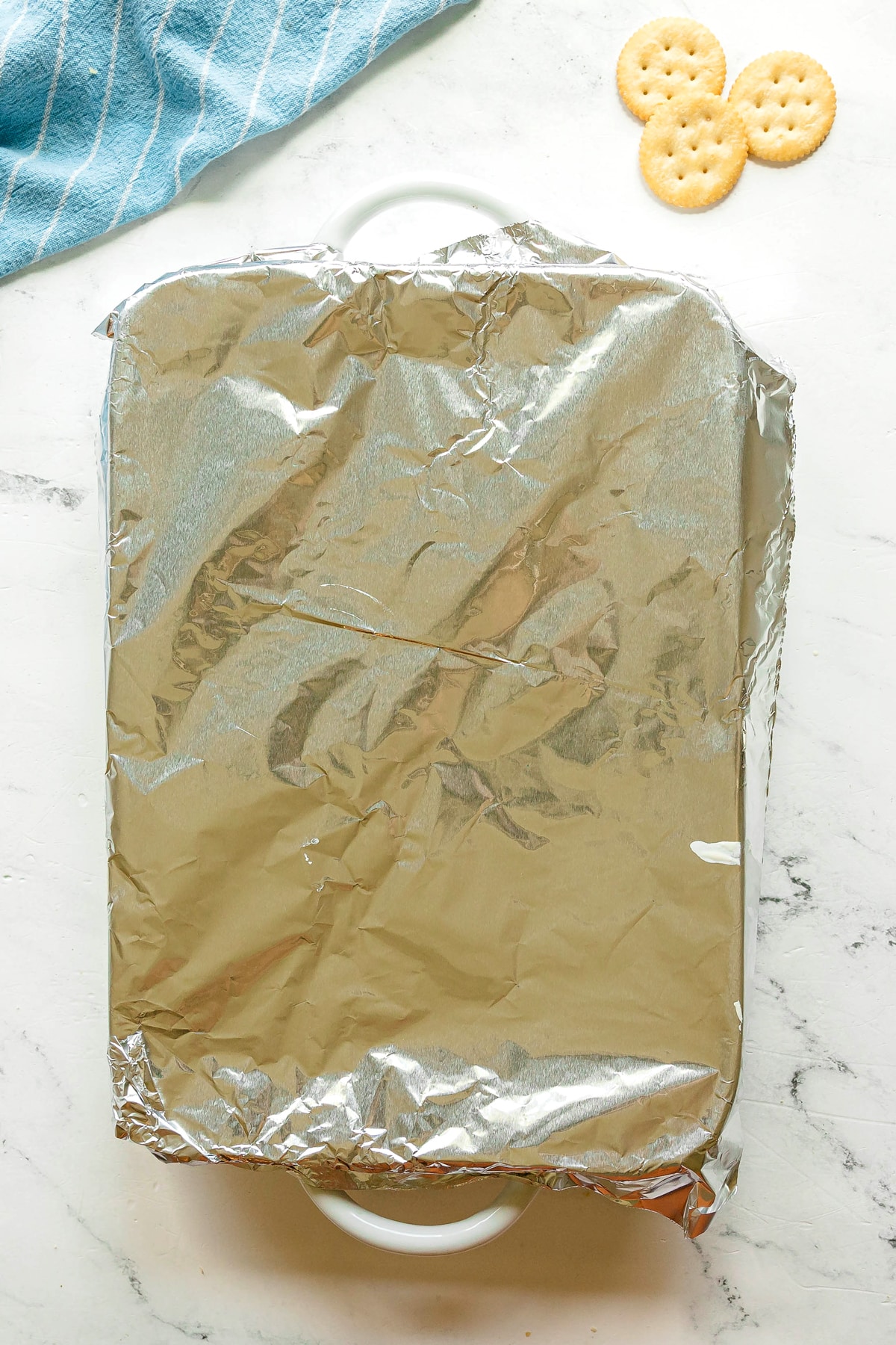 A rectangular baking dish of Ritz Cracker Casserole covered with aluminum foil sits on a white surface next to four round crackers and a blue-striped cloth.