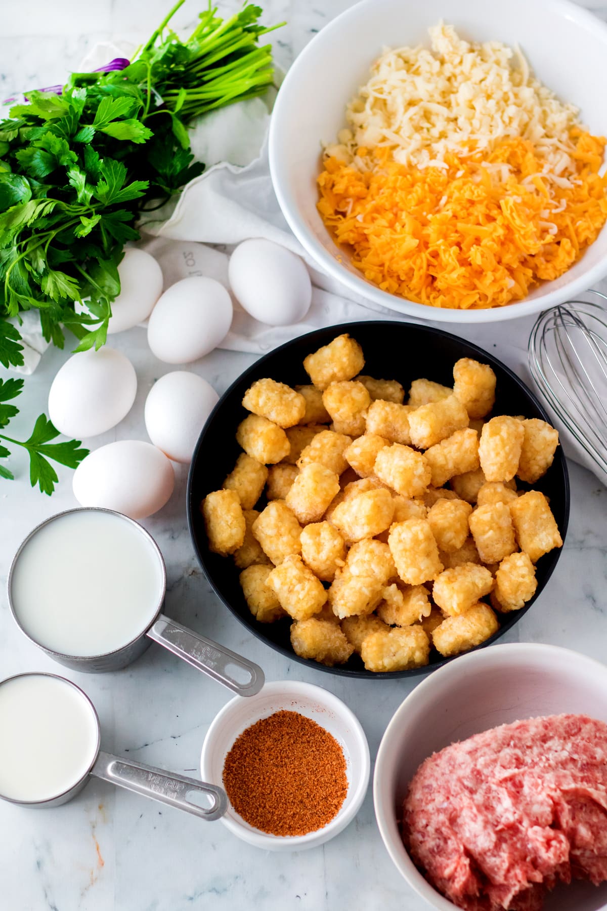Ingredients for Slow Cooker Tator Tot Breakfast Casserole are arranged on a marble countertop. These are: ground breakfast sausage, frozen tater tots, whole milk, cajun seasoning, large eggs, cheddar cheese, pepper jack cheese, and parsley for topping
