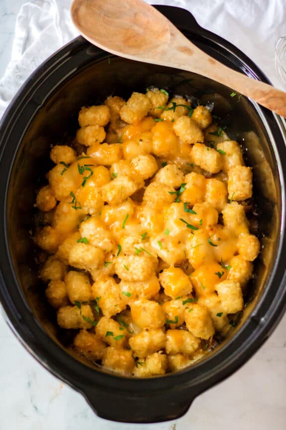 Next step in preparing Slow Cooker Tator Tot Breakfast Casserole is to add melted cheddar cheese and chopped herbs in a black slow cooker, with a wooden spoon resting on the edge.