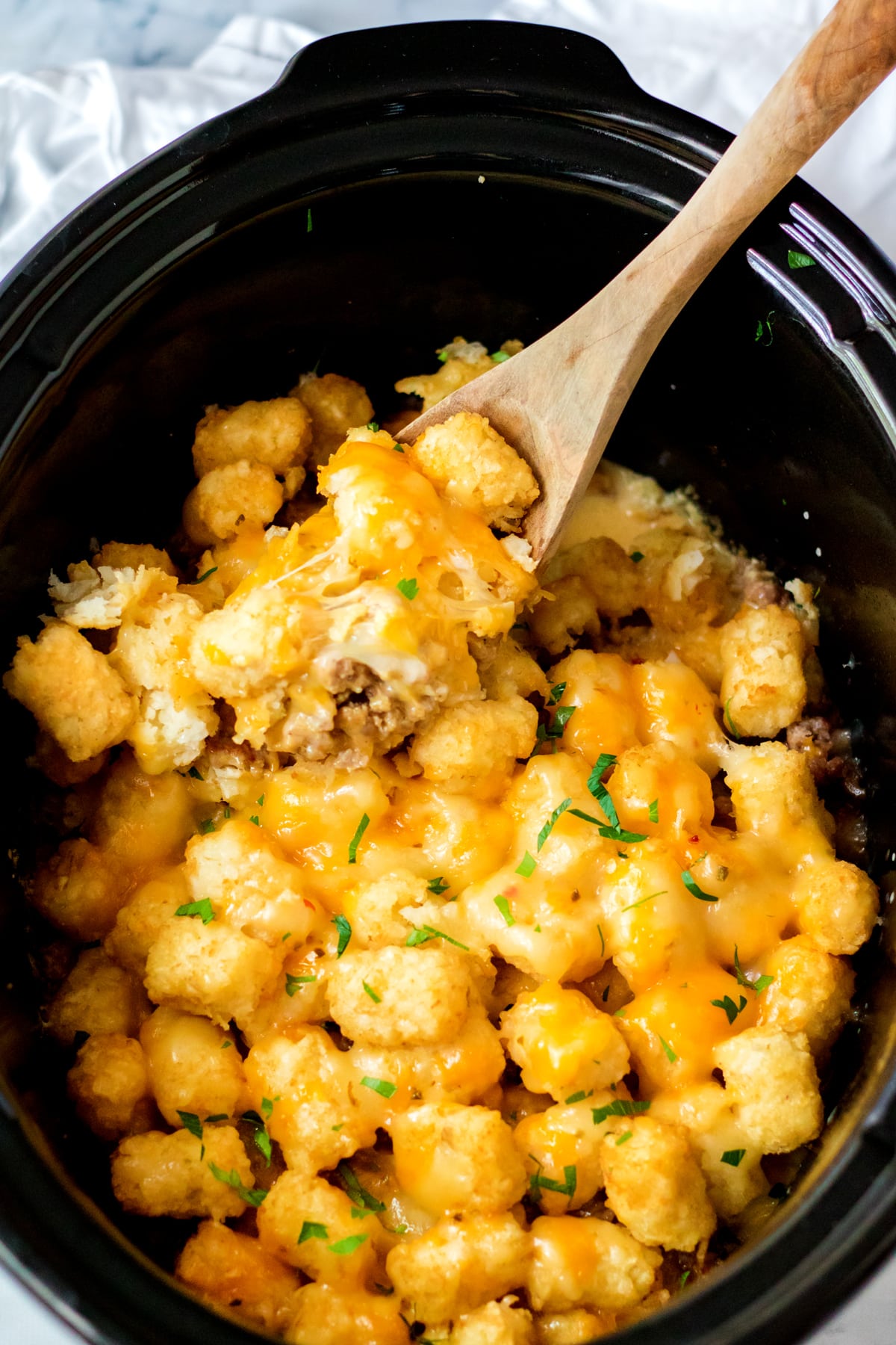A wooden spoon scoops Slow Cooker Tator Tot Breakfast Casserole with ground beef from a black slow cooker, garnished with chopped herbs.