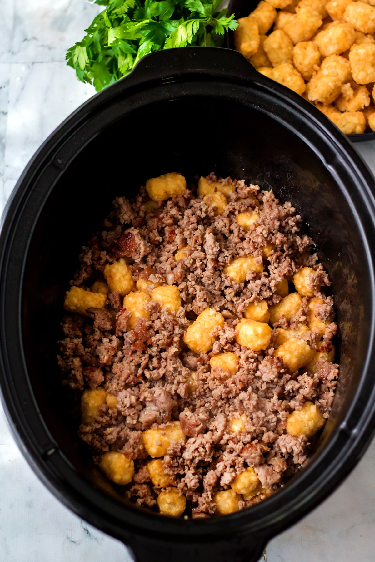 Next process in preparing Slow Cooker Tator Tot Breakfast Casserole is to add ground beef and tater tots layered in a slow cooker. More tater tots and fresh parsley visible in the background.