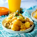 A serving of Slow Cooker Tator Tot Breakfast Casserole with ground meat on a white plate, garnished with chopped herbs, sits on a blue woven cloth. An orange drink and halved orange are in the background.