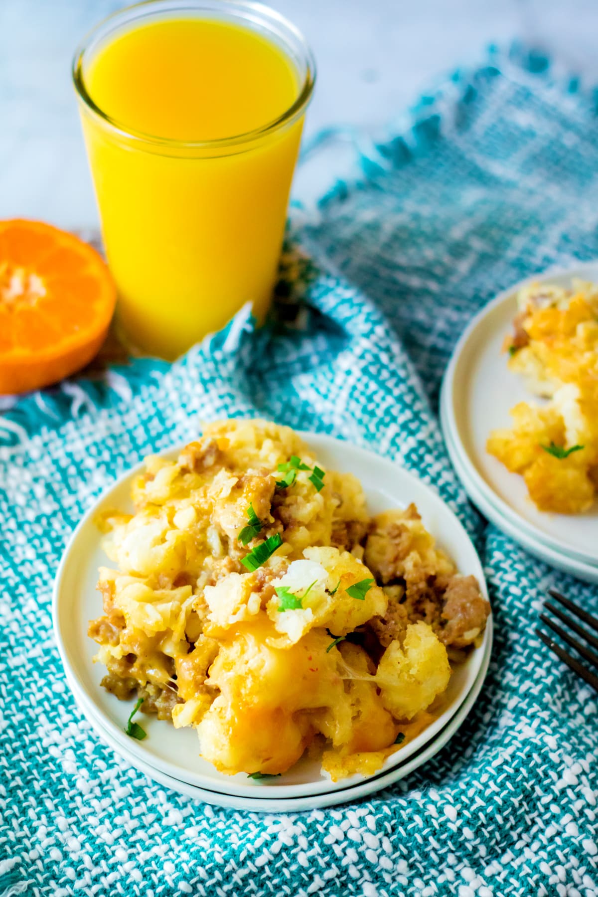 A plate of Slow Cooker Tator Tot Breakfast Casserole with ground meat and potatoes next to a glass of orange juice and a sliced orange on a blue patterned cloth.