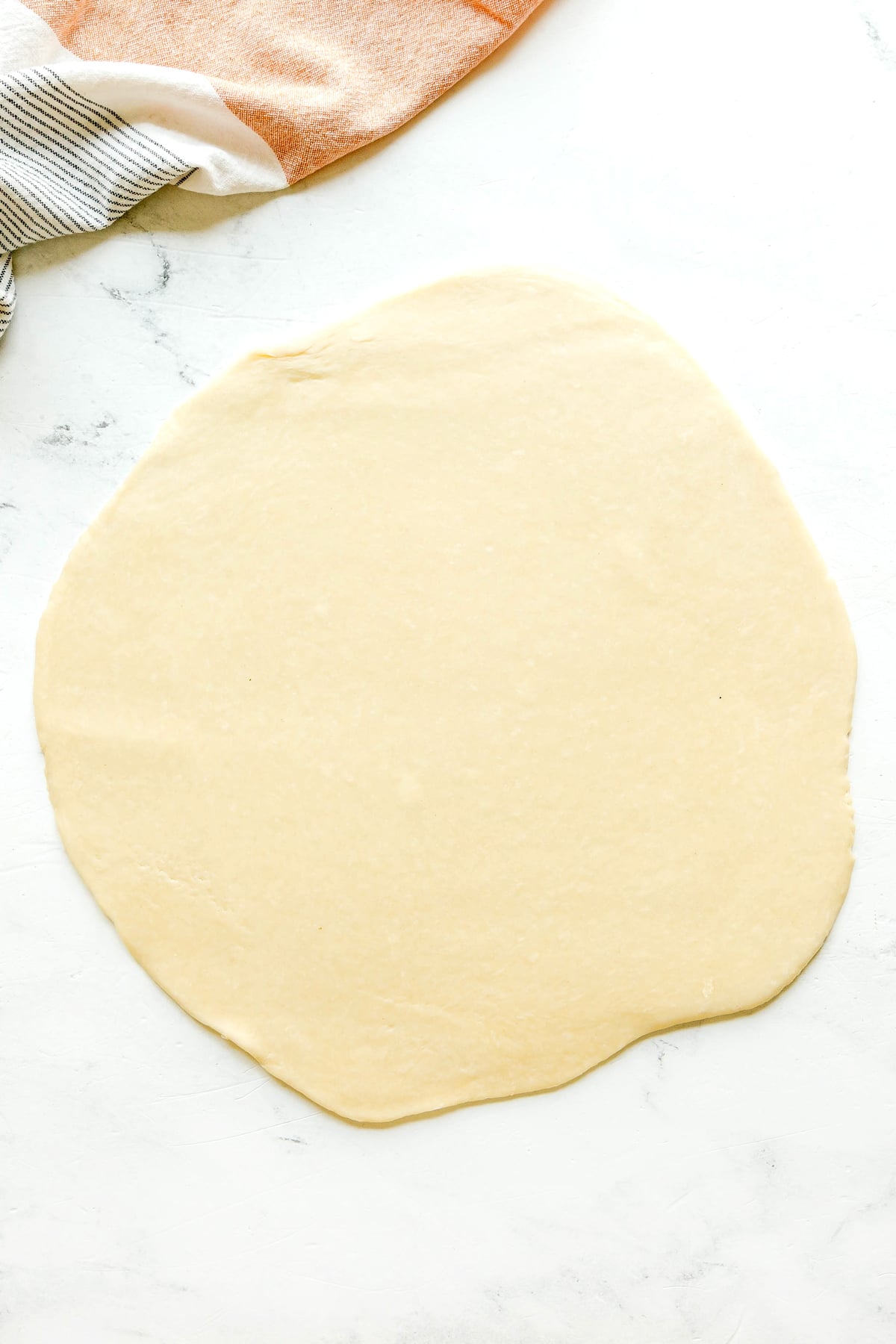 Round sheet of rolled out dough on a white surface for Sweet Potato Pie, with two kitchen towels partially visible in the top left corner.