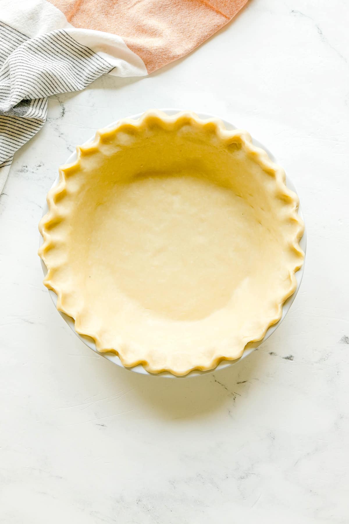 Unbaked pie crust for Sweet Potato Pie with fluted edges in a white pie dish on a light marble surface, with cloths in the background.