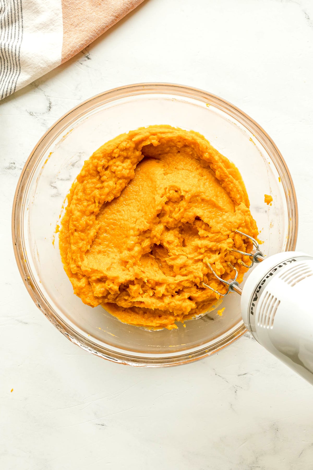 Electric hand mixer blending orange sweet potato mixture in a clear glass bowl on a white surface. A striped kitchen towel is partially visible in the upper left corner. This is a step in preparing Sweet Potato Pie recipe.
