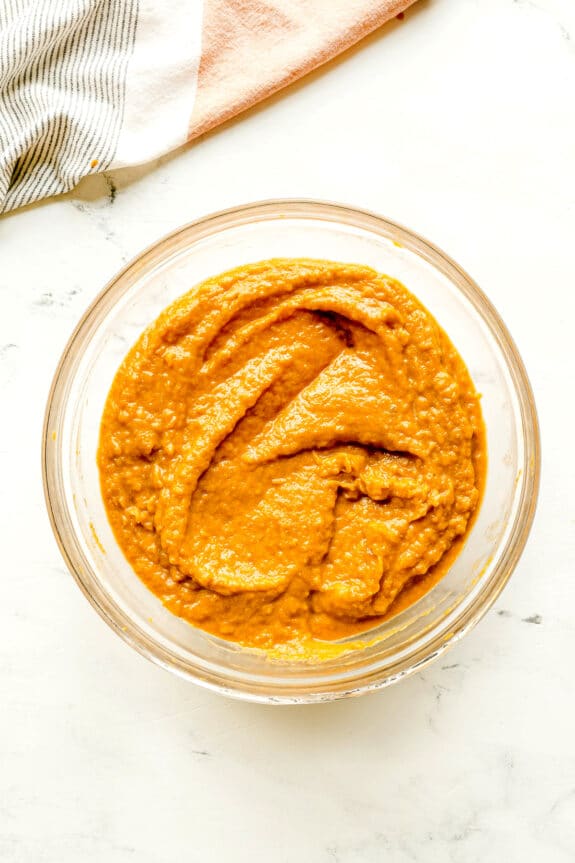 A glass bowl filled with thick, orange-brown puree for Sweet Potato Pie sits on a white surface next to a folded striped cloth.