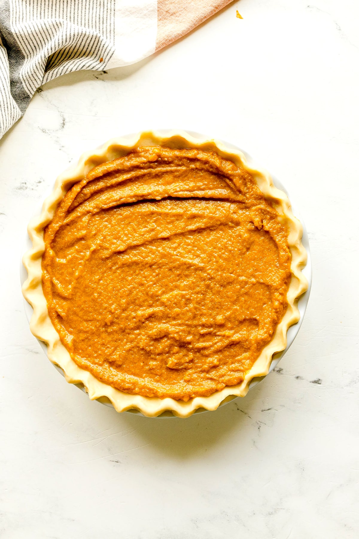 Unbaked Sweet Potato Pie with smooth filling in a fluted crust, placed on a white surface next to a striped cloth.