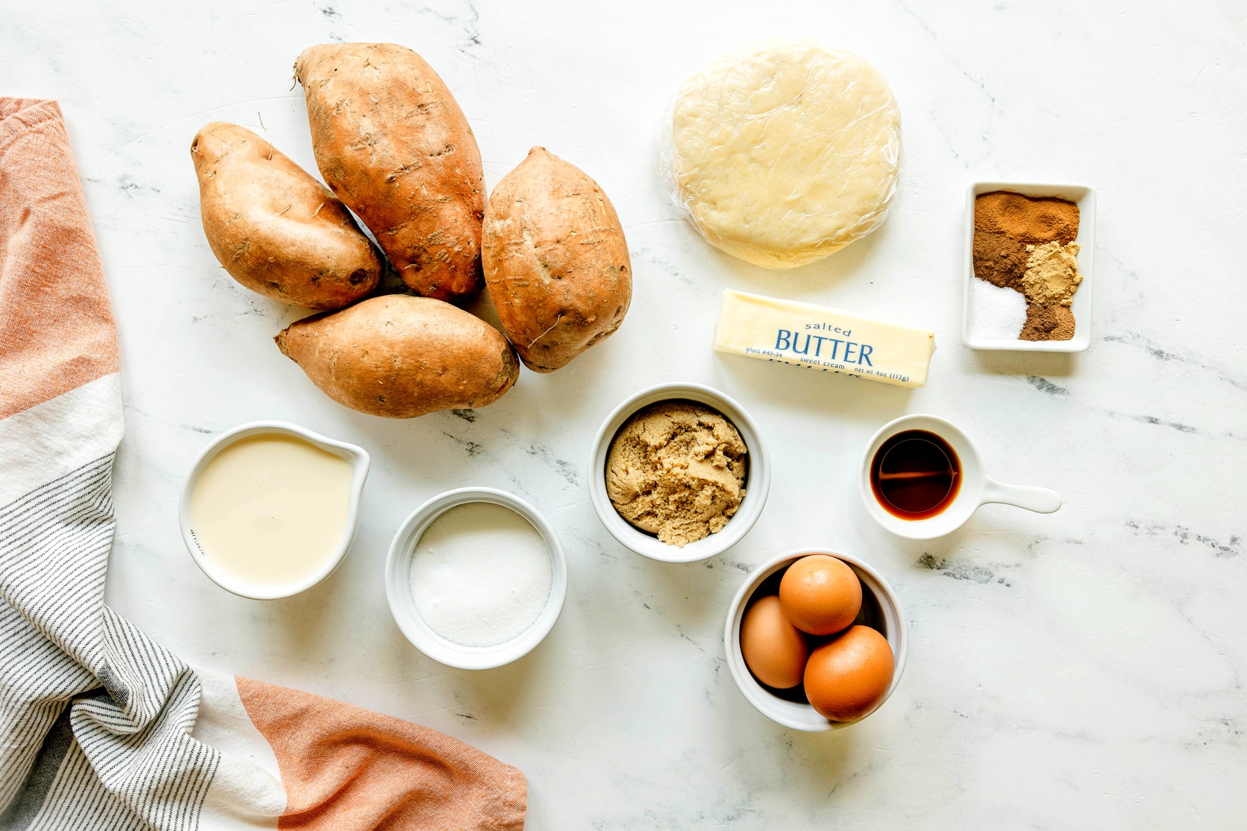 Overhead view of sweet potatoes, pie crust, eggs, butter, brown and white sugar, vanilla, evaporated milk, and spices on a marble surface with a kitchen towel.