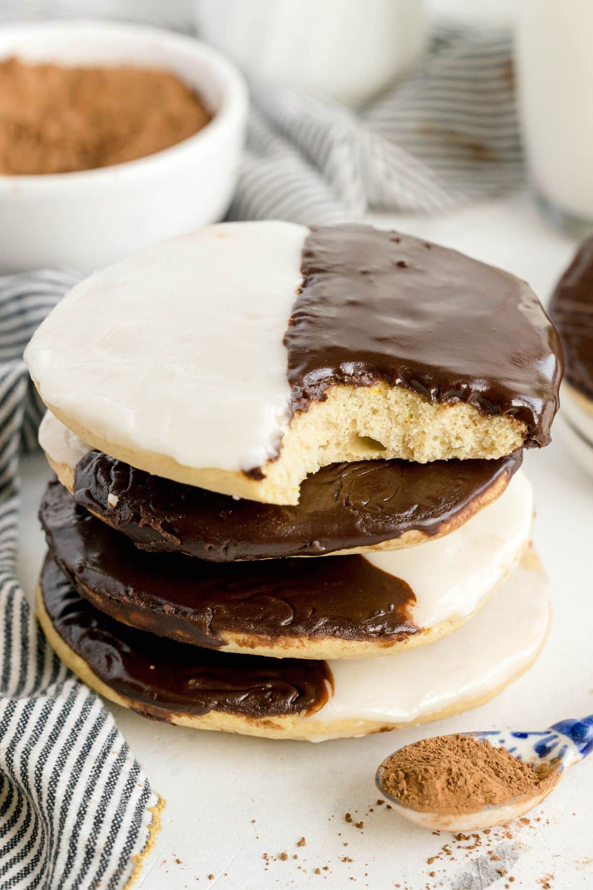 A stack of black and white cookies, made from a classic black and white cookies recipe, sits on a white surface with the top cookie partially eaten, next to a striped cloth and a wooden spoon sprinkled with cocoa powder.