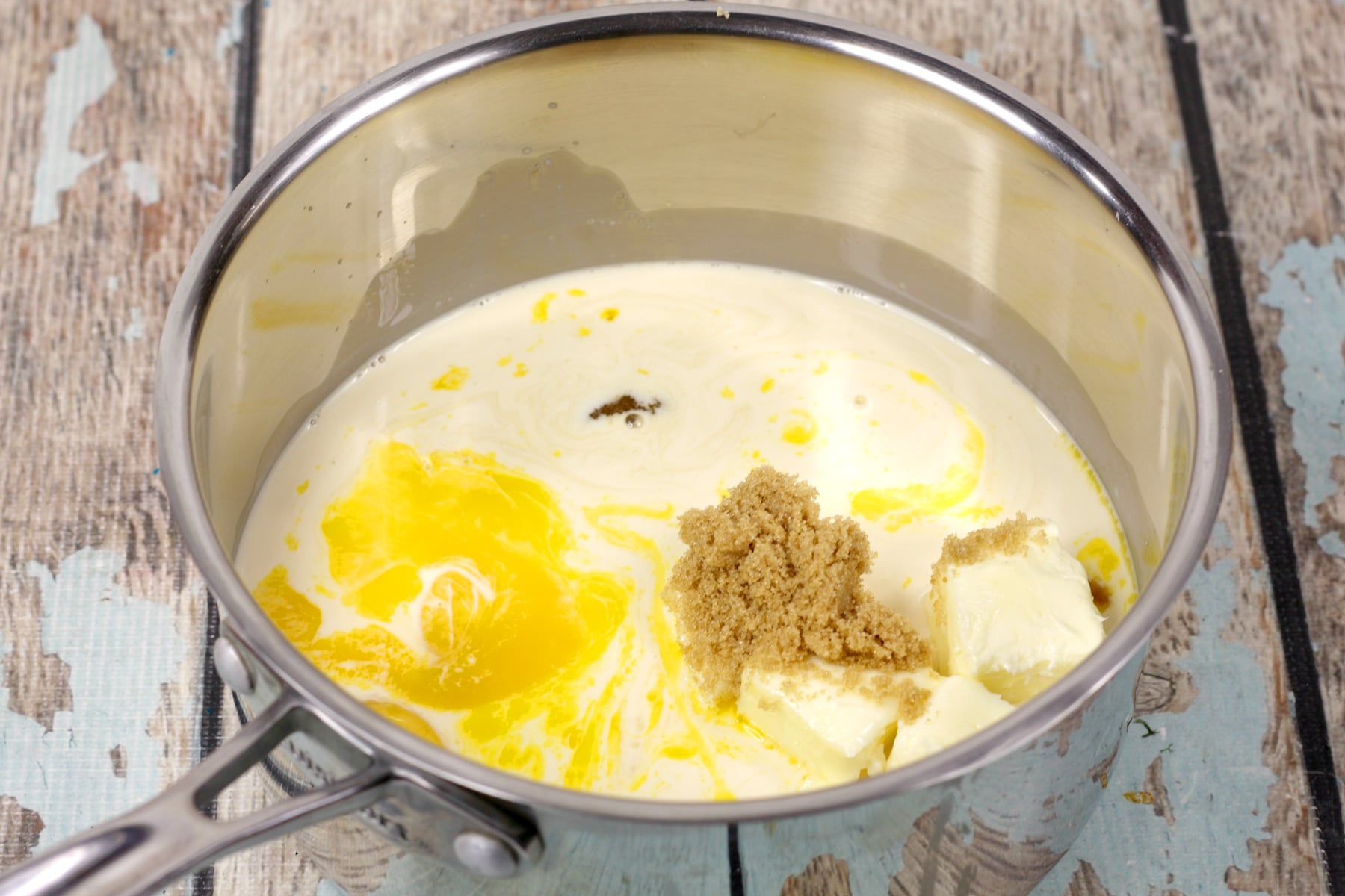 A saucepan on a rustic wooden surface contains milk, melted butter, brown sugar, and vanilla, ready to be mixed for German Chocolate Brownies recipe