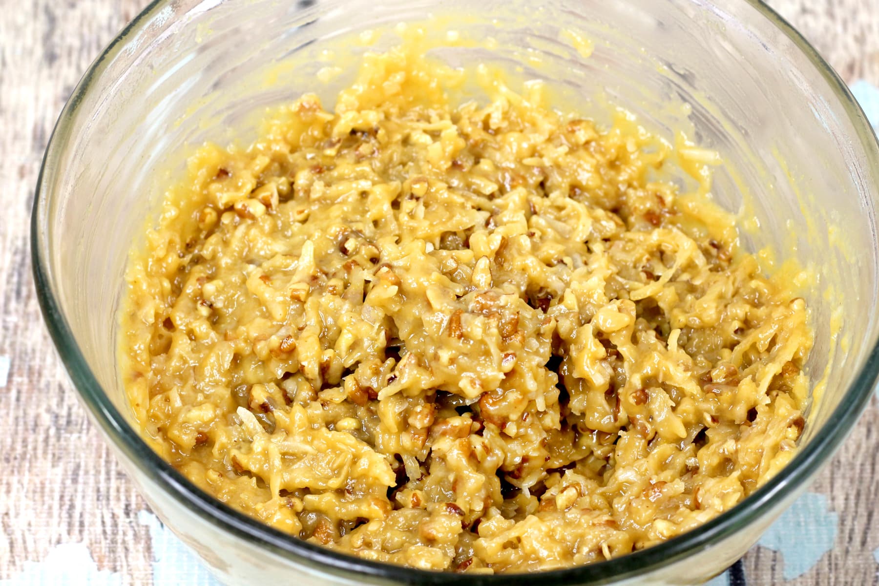 A clear glass bowl filled with a thick, chunky yellow mixture containing shredded coconut and nuts sits on a wooden surface for German Chocolate Brownies recipe.
