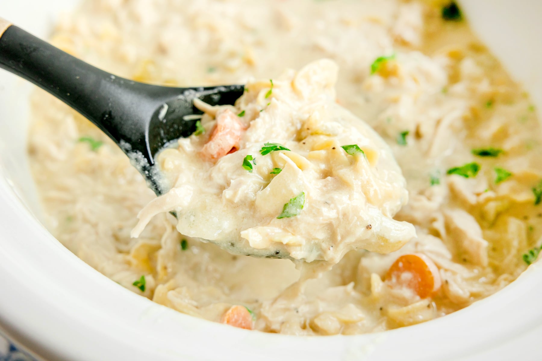 A black spoon lifts a serving of Slow Cooker Chicken and Noodles with visible chunks of chicken, carrots, and herbs from a white bowl.