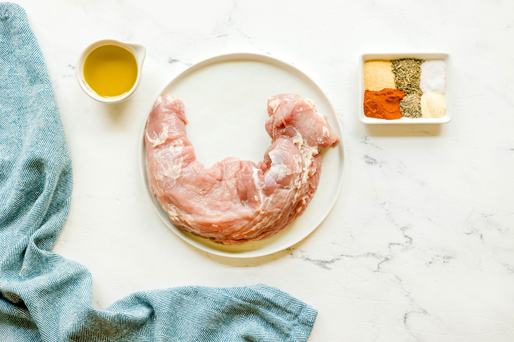 Ingredients for Air Fryer Pork Tenderloin are the following: pork tenderloin, olive oil, garlic powder, smoked paprika, Italian seasoning, kosher salt, onion powder, and black pepper, with blue linen on the side.