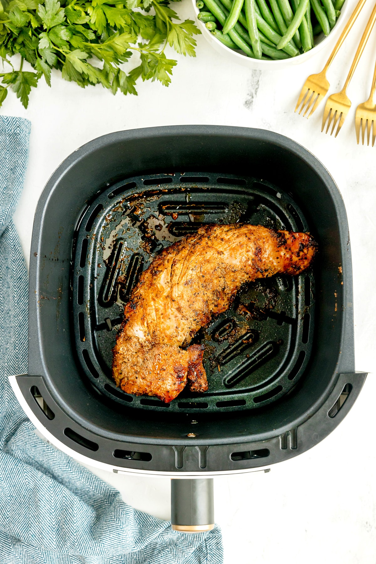 A piece of Air Fryer Pork Tenderloin meat after cooking at 400°F for 18 to 22 minutes. Linen cloth can be seen on the side with utensils , beans, and parsley leaves.