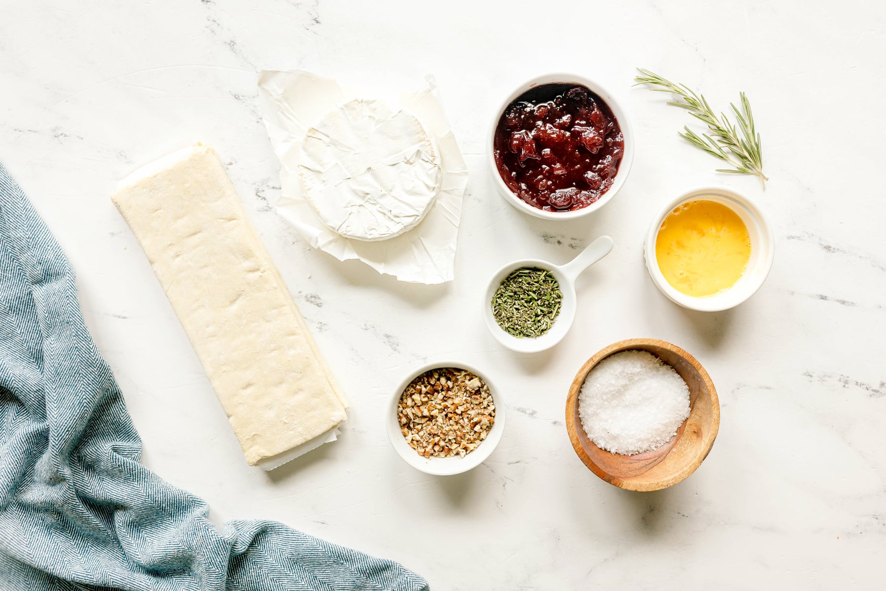 Ingredients for Cranberry Brie Bites include a round of brie, frozen puff pastry, cranberry sauce, chopped pecans, finely chopped rosemary, and sea salt on a white surface with a blue cloth.