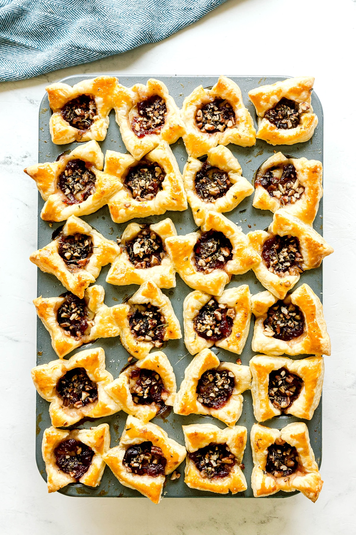 A muffin tin filled with 24 square-shaped Cranberry Brie Bites, each with a fruit filling and chopped nuts in the center, sits on a white surface with a blue cloth in the background.