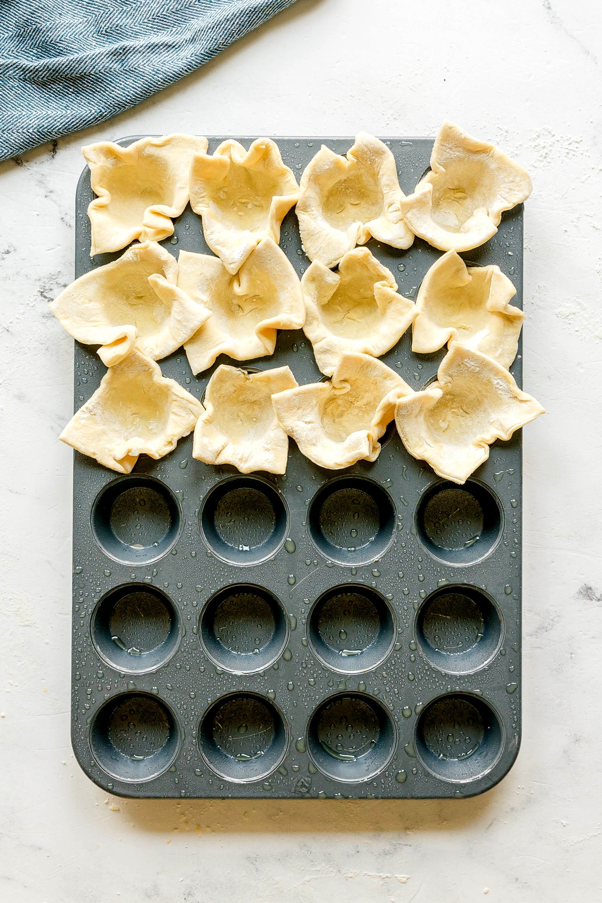 A muffin tin is partially filled with squares of dough pressed into the cups, with eight cups remaining empty. A blue cloth is in the top left corner. This is one of the steps in preparing Cranberry Brie Bites.