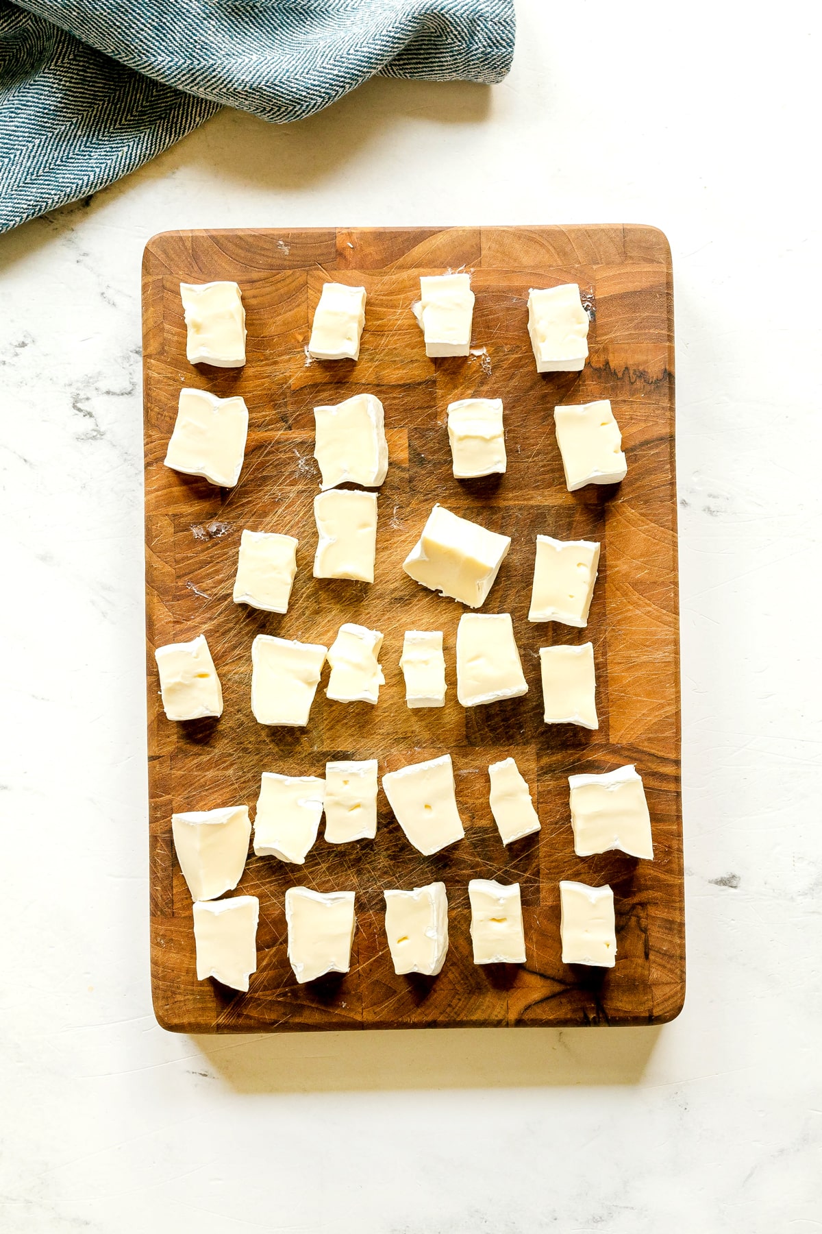 Next step in preparing Cranberry Brie Bites is to cut 25 evenly spaced cubes of brie on a white marble surface, next to a folded blue cloth.