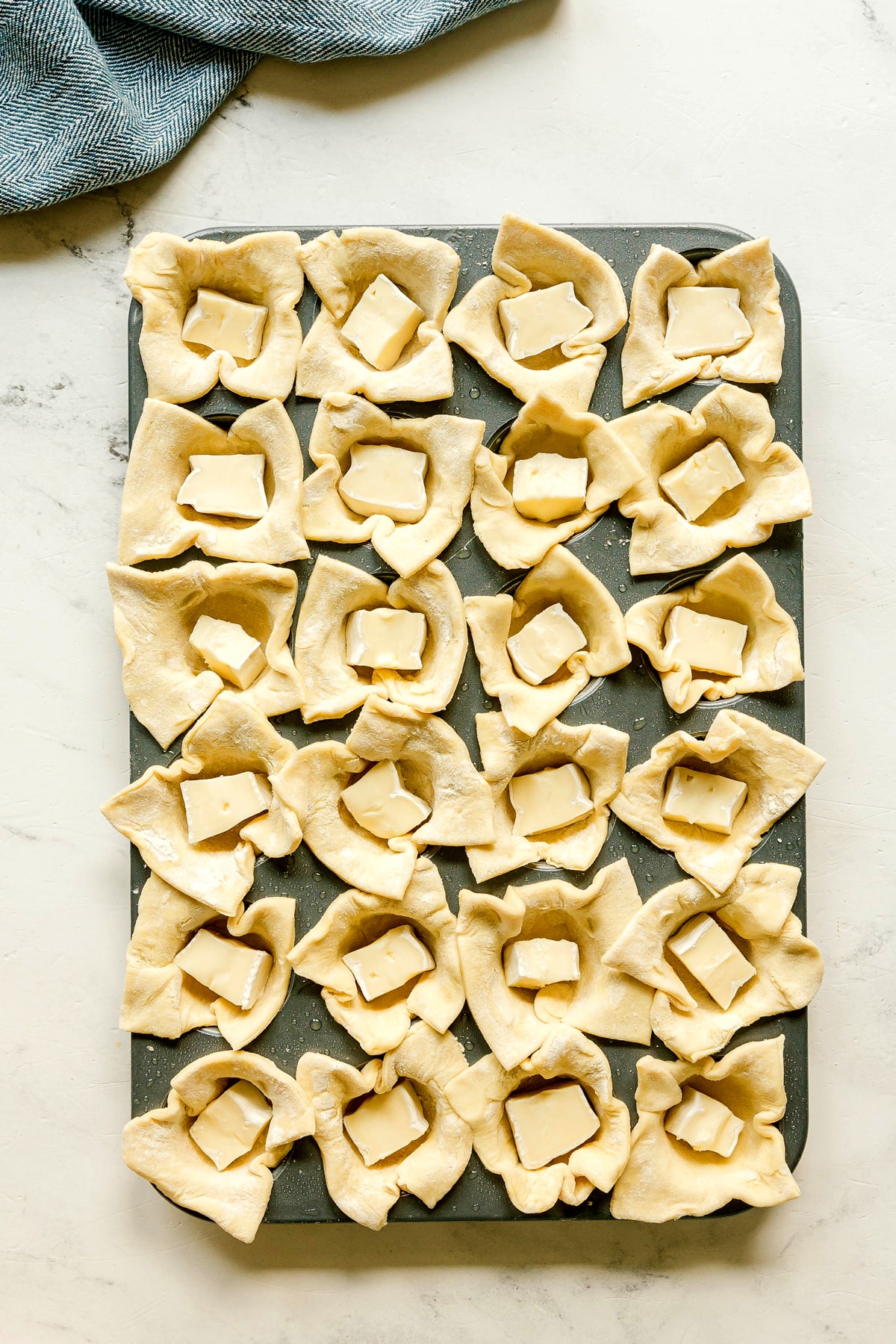 Muffin tin filled with unbaked pastry dough squares, each with a piece of brie in the center, ready to be baked as one process in preparing Cranberry Brie Bites