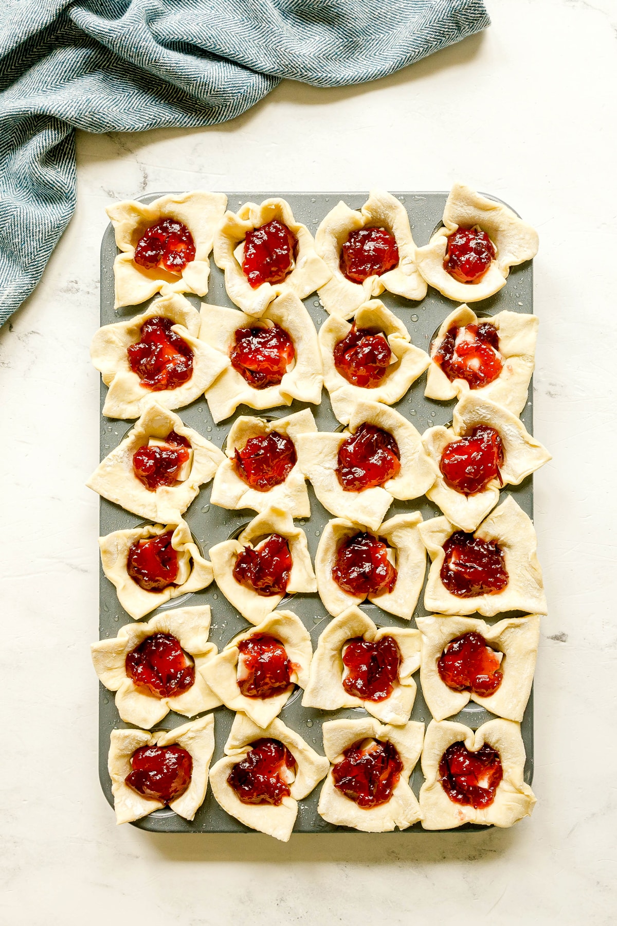 A baking tray holds 24 pieces of unbaked pastry dough folded around a spoonful of cranberry sauce, arranged in a grid for Cranberry Brie Bites recipe. A blue and white cloth is partially visible in the upper left corner.