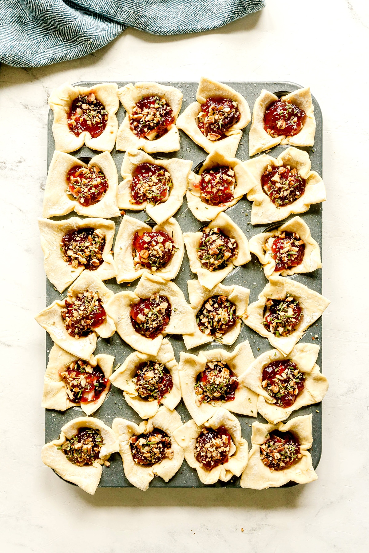 A baking tray filled with 24 uncooked Cranberry Brie Bites, each topped with cranberry sauce, chopped pecans and rosemary, sits on a white surface.