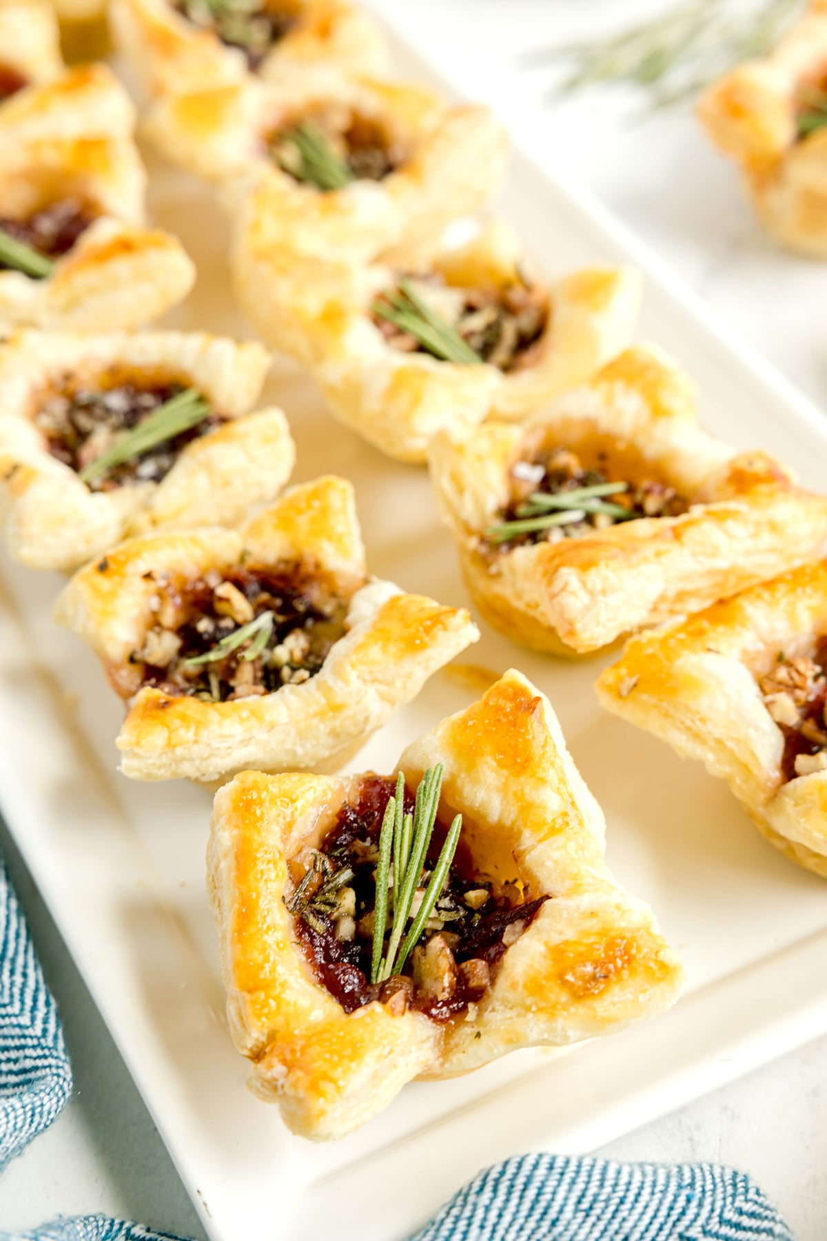 Cranberry Brie Bites filled with cranberry sauce, nuts, and topped with rosemary sprigs are arranged neatly on a rectangular white plate.