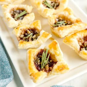 Rectangular white plate with several Cranberry Brie Bites filled with a cranberry sauce, chopped nuts, and garnished with small sprigs of rosemary.
