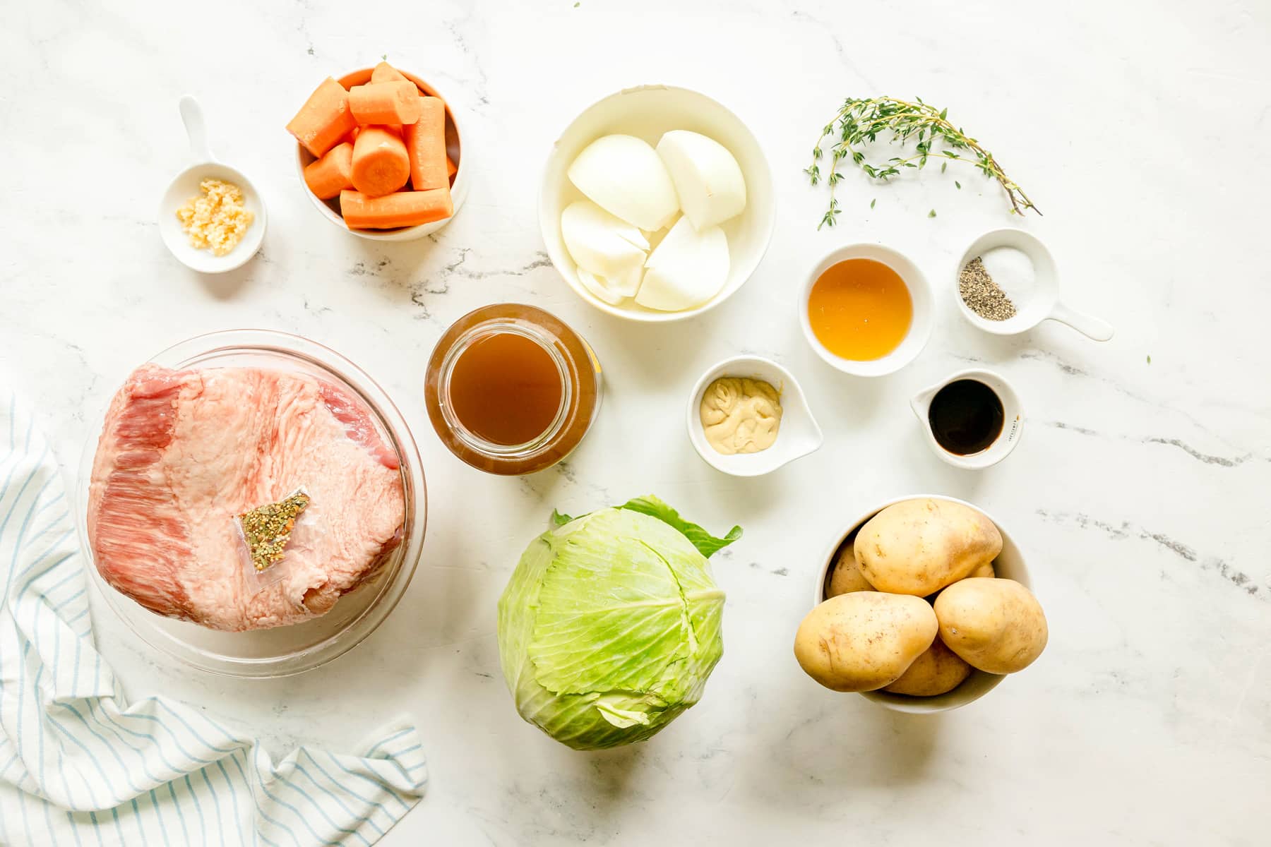 Top-down view of ingredients for Crockpot Corned Beef and Cabbage: carrots, onion, garlic, fresh thyme, corned beef brisket, beef broth, worcestershire sauce, teaspoon salt, black pepper, green cabbage, yukon gold potatoes, dijon mustard, and honey, arrange on a white surface.