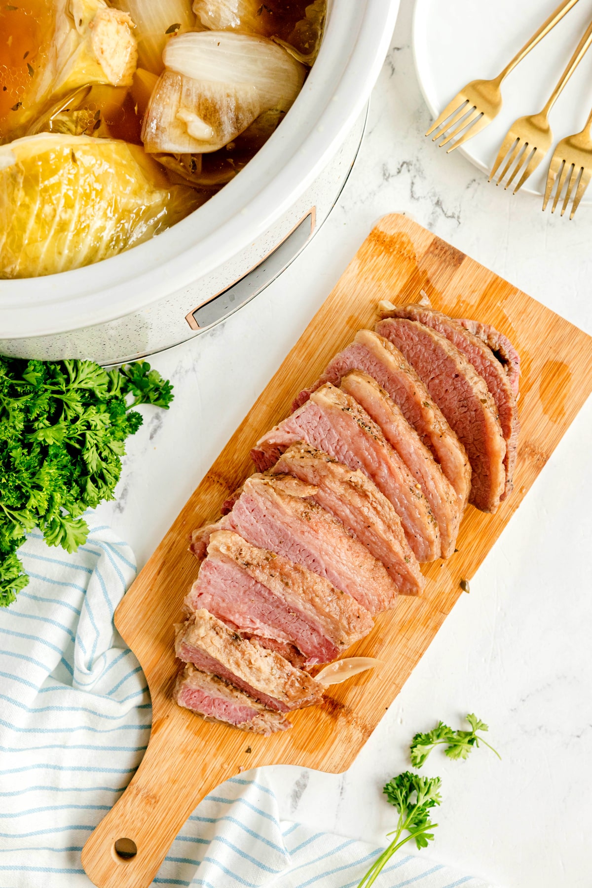Sliced corned beef on a wooden cutting board next to a slow cooker with cabbage and plates with gold forks. This is one of the steps in preparing Crockpot Corned Beef and Cabbage recipe.