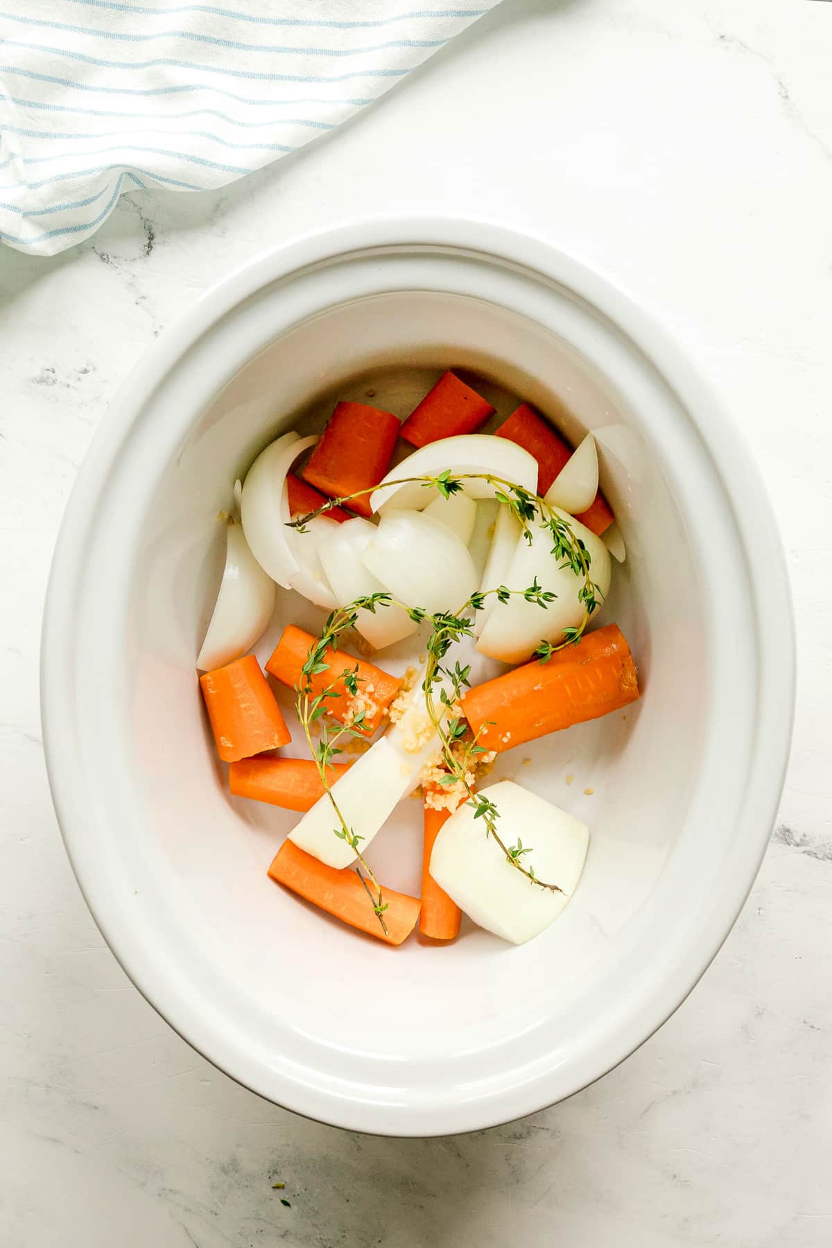 First step in preparing Crockpot Corned Beef and Cabbage is to add chopped carrots, onions, garlic, and sprigs of thyme on a white slow cooker on a white surface.