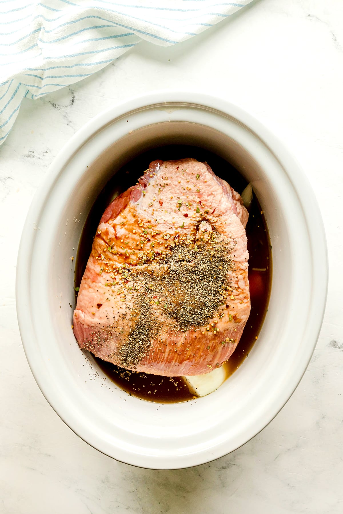 Raw seasoned beef brisket for Crockpot Corned Beef and Cabbage recipe, in a slow cooker with seasonings broth and sauce, ready to cook; white background with part of a striped cloth visible in the corner.