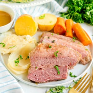 A plate of Crockpot Corned Beef and Cabbage with sliced corned beef, boiled potatoes, carrots, cabbage, and a side of brown sauce, garnished with parsley.
