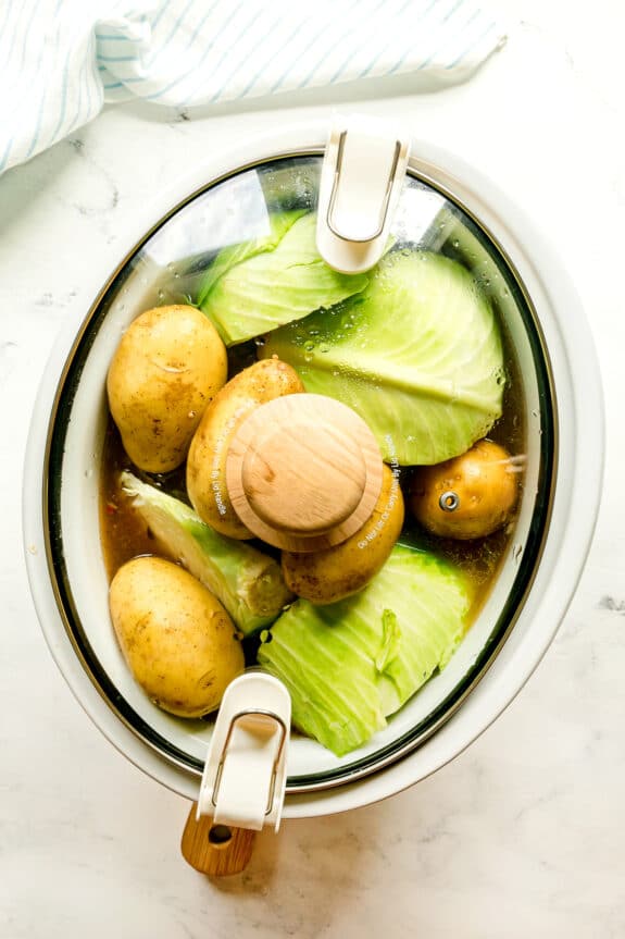 Next step in preparing Crockpot Corned Beef and Cabbage is to add whole potatoes and cabbage wedges to the beef brisket, then cover it with glass lid.