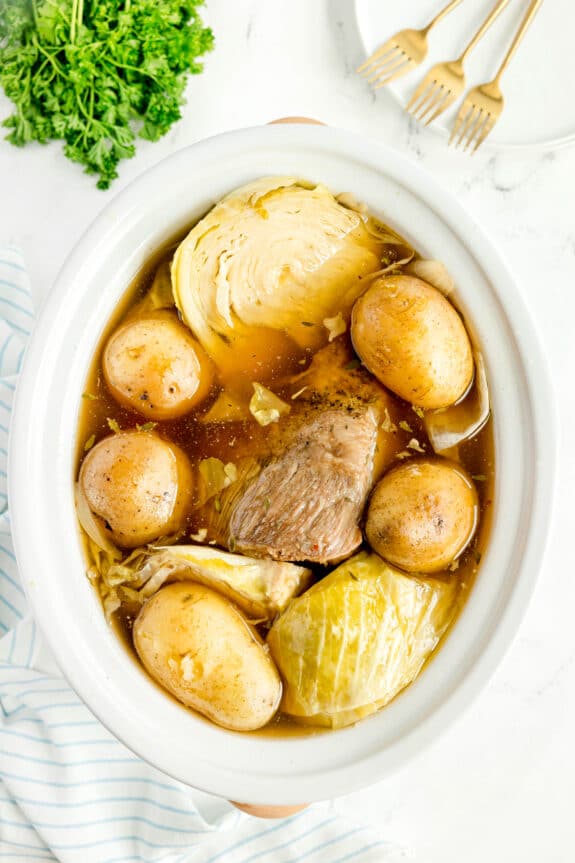 Oval dish with cooked beef, whole potatoes, cabbage wedges, and broth or Crockpot Corned Beef and Cabbage recipe. Plates, forks, parsley, and a striped cloth are nearby on a white surface.
