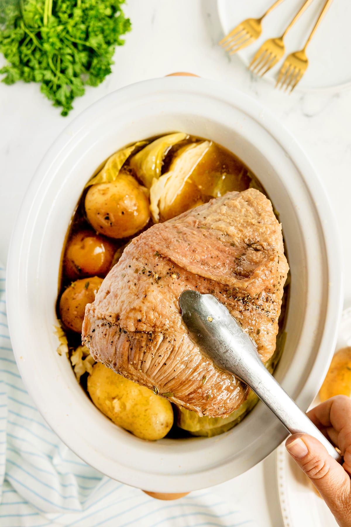 A hand uses tongs to lift cooked corned beef from a slow cooker with potatoes and cabbage. Parsley and gold forks are visible nearby. This is one step in preparing Crockpot Corned Beef and Cabbage recipe.