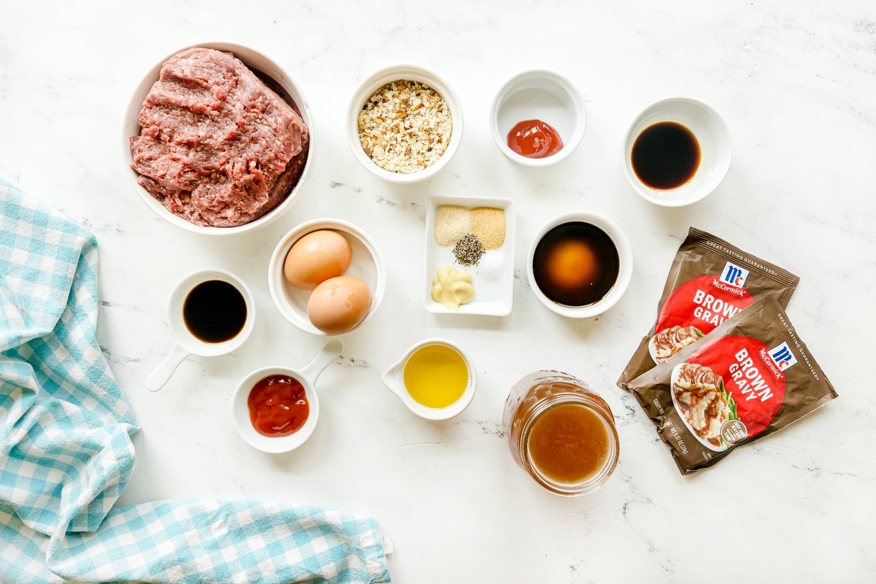 Various ingredients for Crockpot Salisbury Steak include ground beef, breadcrumbs, eggs, worcestershire sauce, ketchup, dijon mustard, garlic powder, onion powder, salt and black pepper and olive oil on a white surface beside a blue checkered cloth.