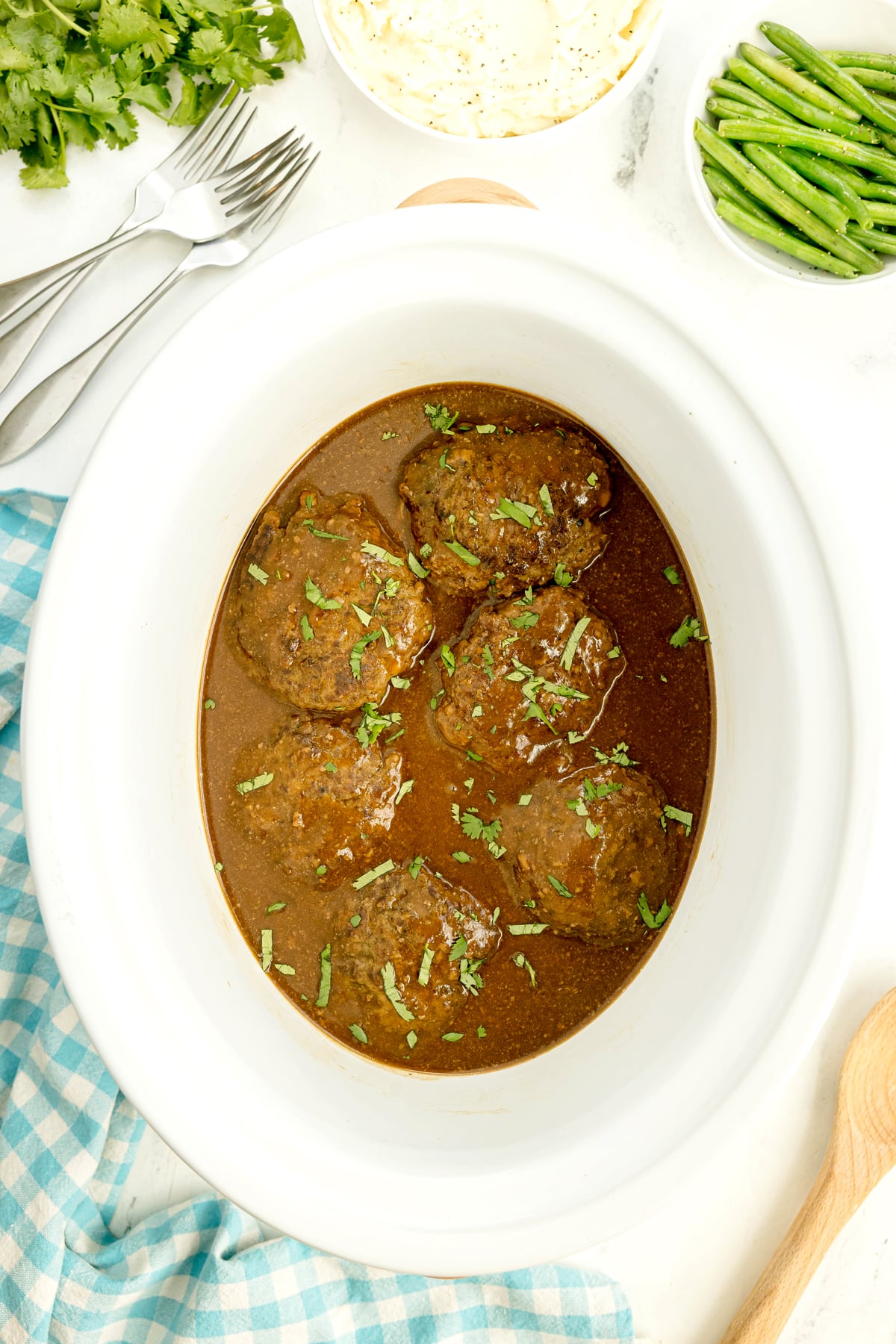 Six beef patties of Crockpot Salisbury Steak sit in a white slow cooker, garnished with chopped herbs. Green beans, cilantro, and forks are nearby on the table.