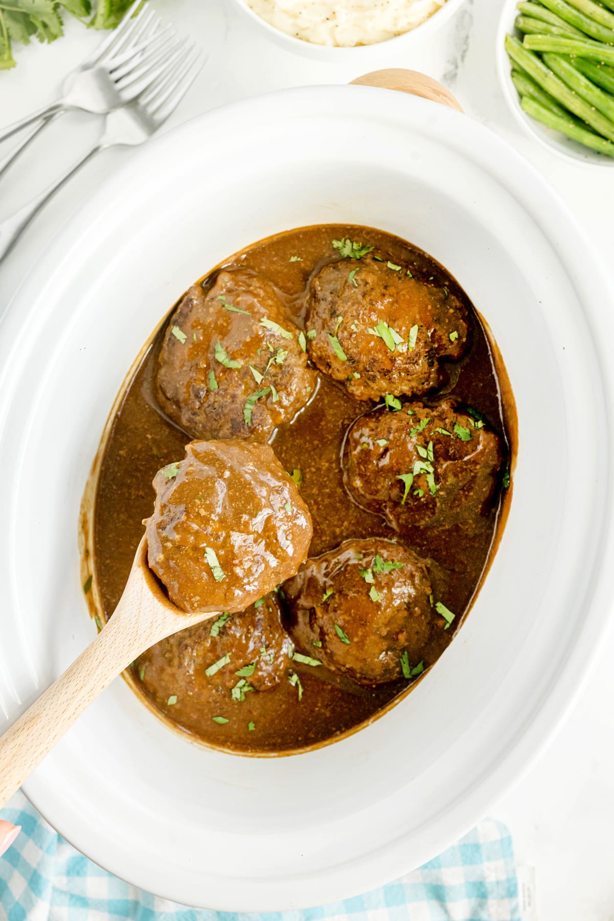 Crockpot Salisbury Steak with five meatballs in brown gravy are served in a white oval slow cooker, garnished with chopped herbs, with a wooden spoon lifting one meatball.