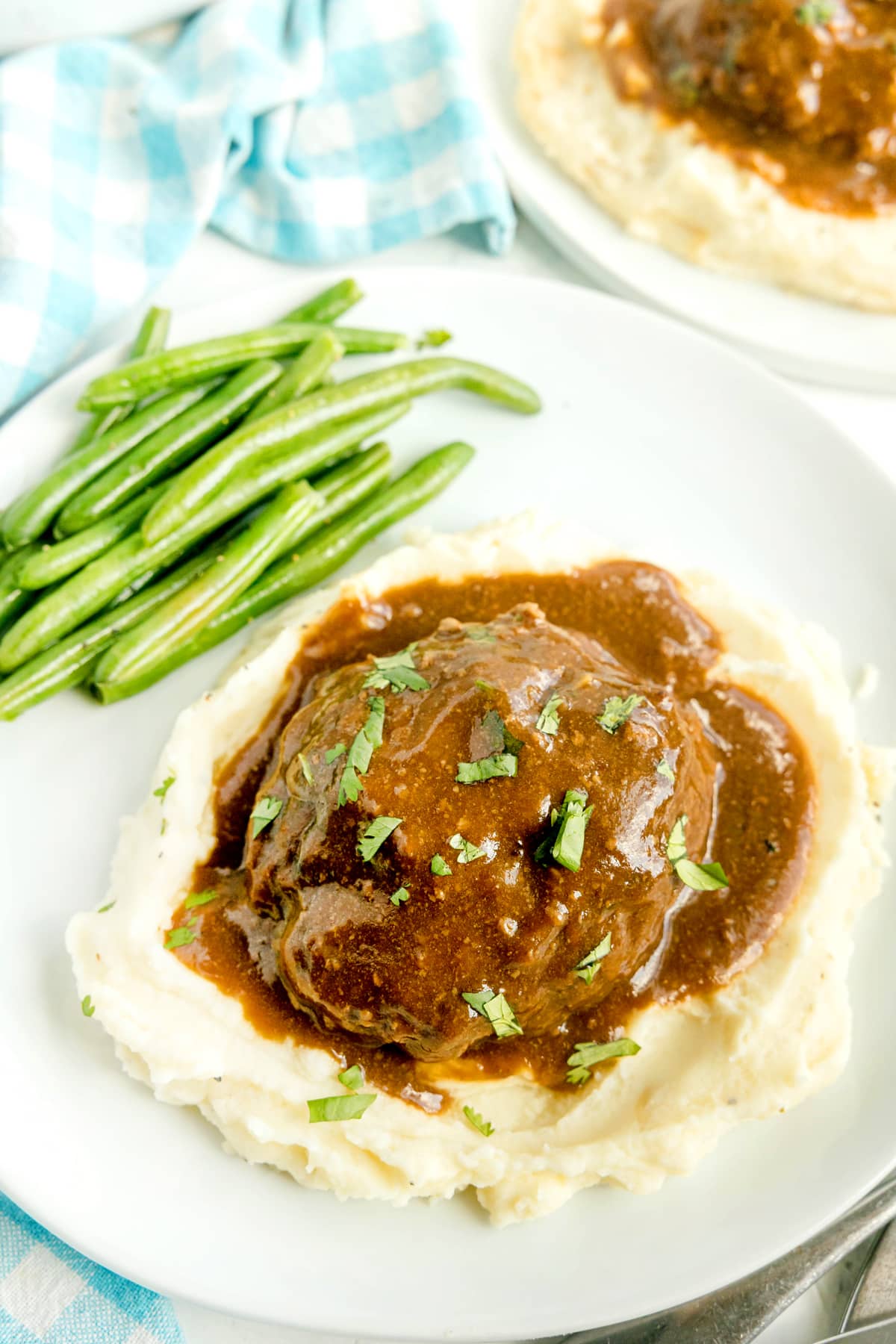 A plate of Crockpot Salisbury Steak with mashed potatoes topped with brown gravy and herbs, served alongside cooked green beans on a white dish.