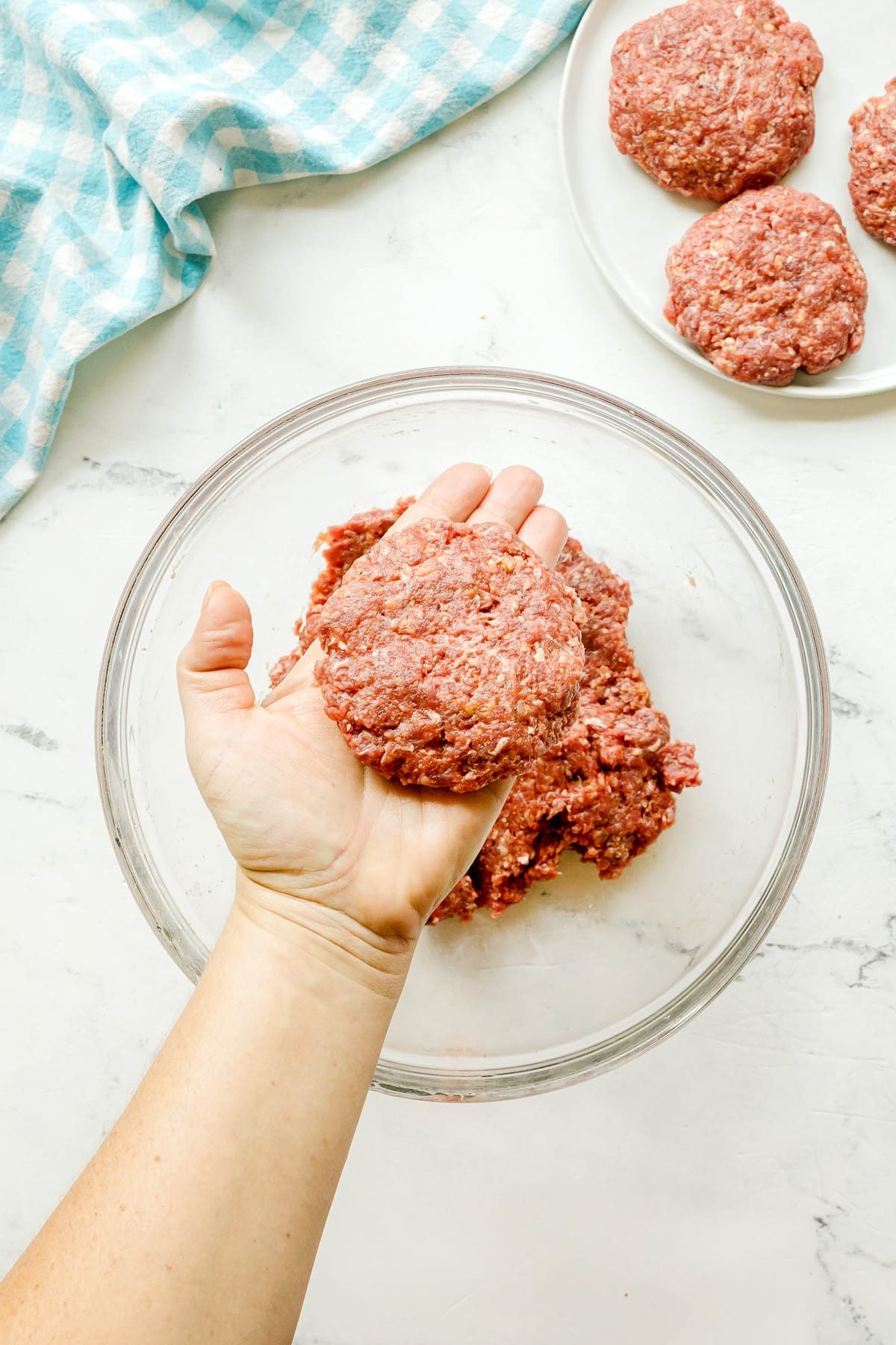A hand holds a raw hamburger patty for Crockpot Salisbury Steak recipe over a glass bowl of ground meat, with more patties on a plate and a blue checked cloth nearby.