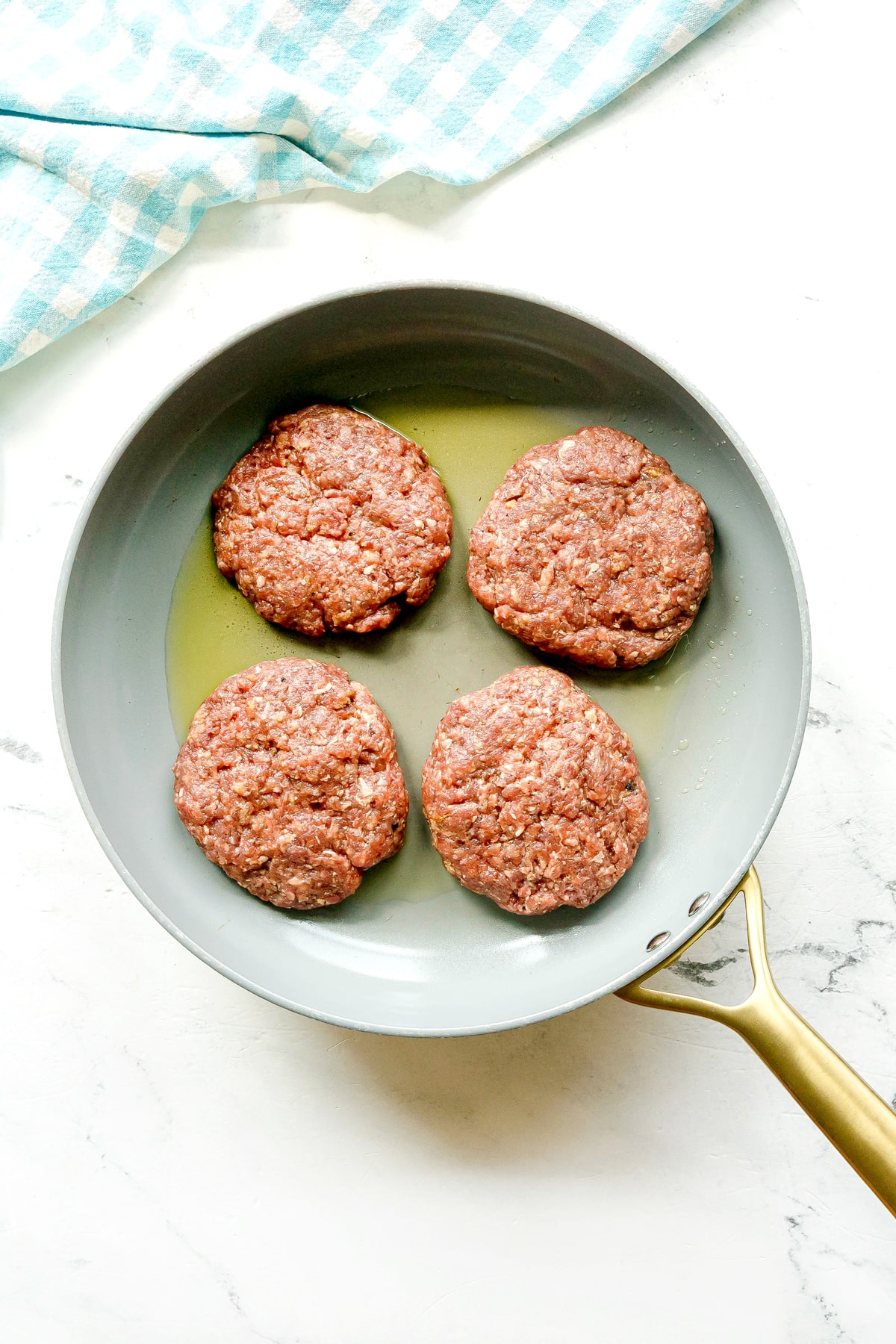 Four raw hamburger patties for Crockpot Salisbury Steak are cooking in a frying pan with some oil. The pan is on a white surface next to a blue and white checkered cloth.