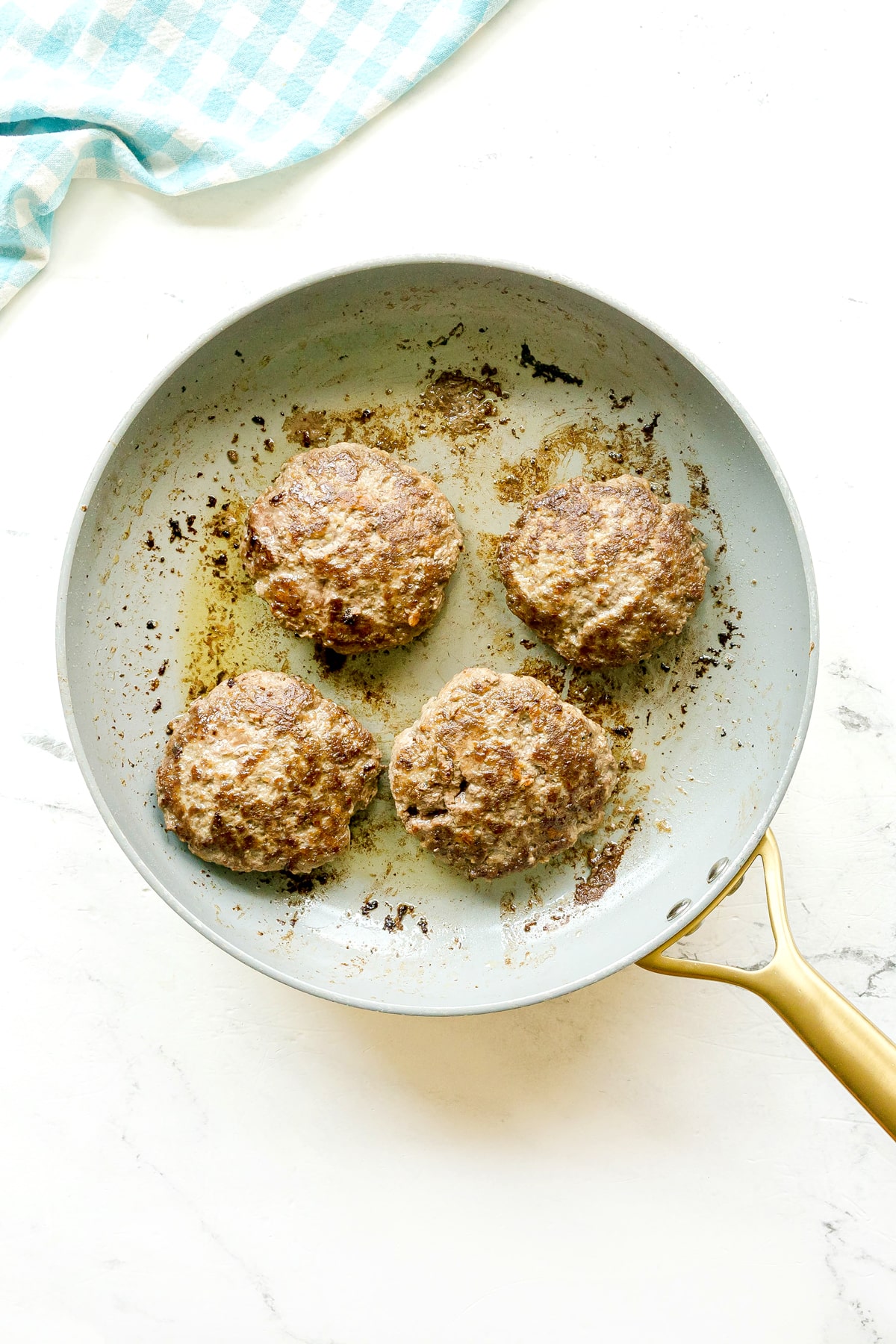 Four cooked beef patties for Crockpot Salisbury Steak recipe in a light-colored frying pan with a gold handle, viewed from above, next to a blue and white checkered cloth.