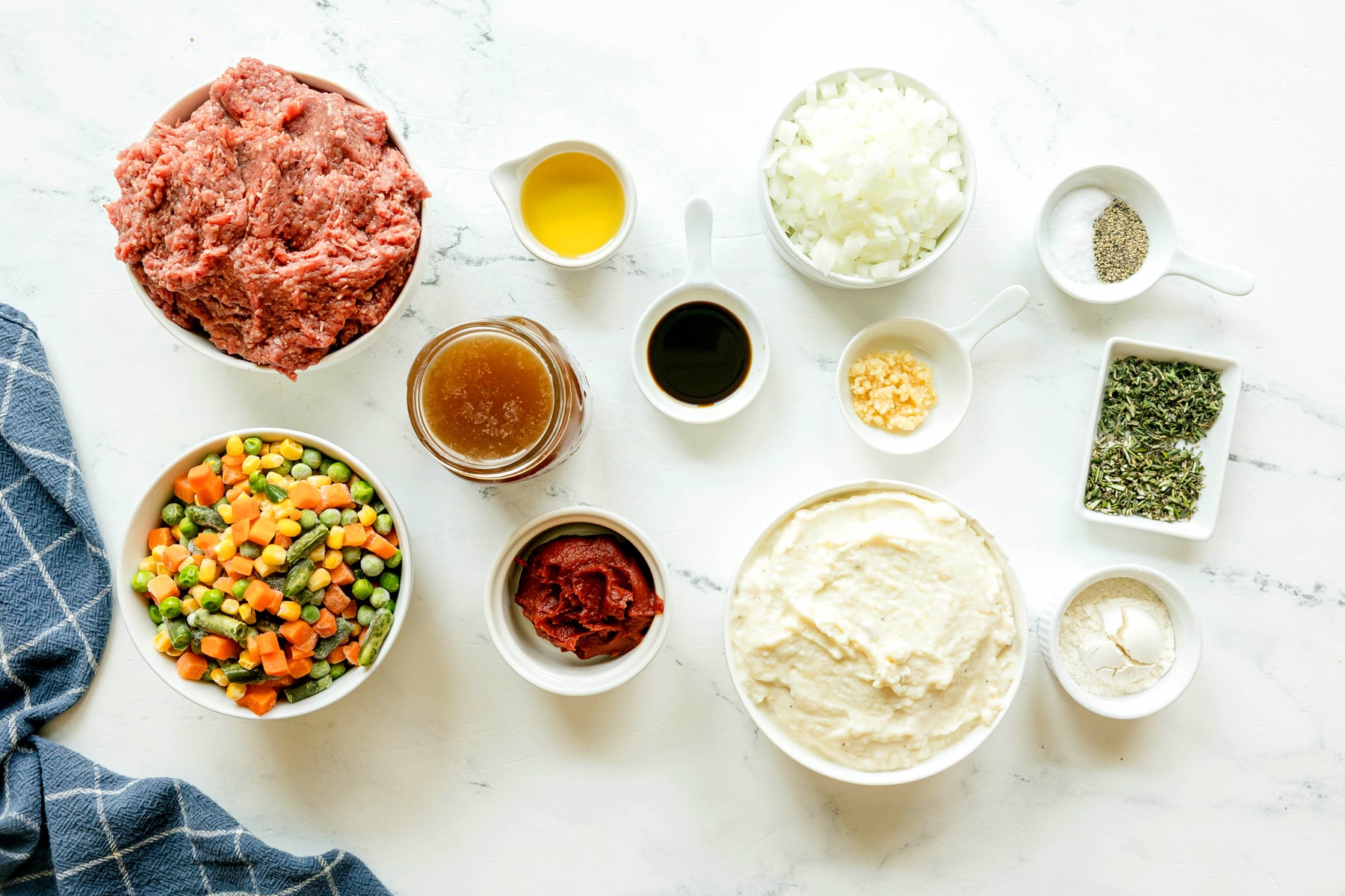 Various ingredients for Crockpot Shepherd's Pie recipe, including ground beef, vegetable oil, onion, tomato paste, garlic, fresh rosemary and thyme, salt and black pepper, worcestershire sauce, beef stock, flour, frozen mixed vegetable and mashed potatoes and a blue towel on side.