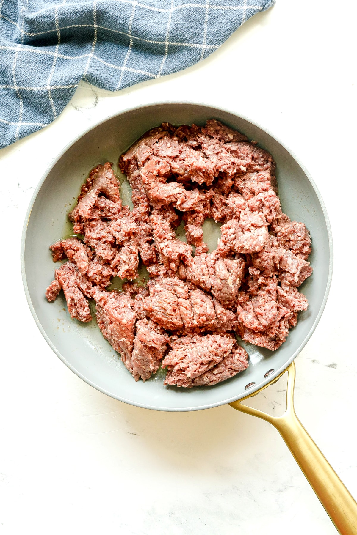 First step in preparing Crockpot Shepherd's Pie is to brown the beef in a frying pan on a white surface. A blue and white checkered cloth cab be seen nearby.