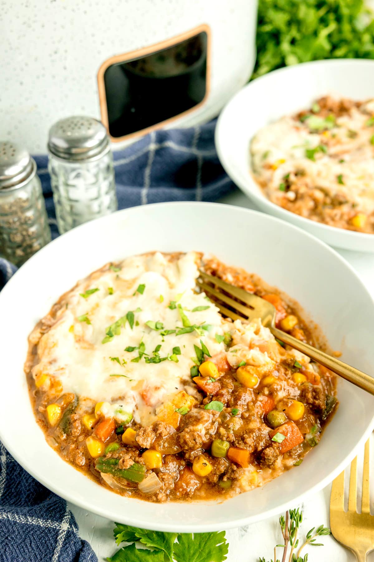 A plate of Crockpot Shepherd's Pie with ground meat, mixed vegetables, and mashed potatoes, garnished with chopped herbs, sits on a blue napkin next to salt and pepper shakers.